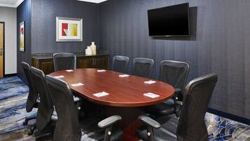 A conference room with a large oval table, six chairs, a flat-screen TV mounted on the wall, and a decorative vase on a cabinet.