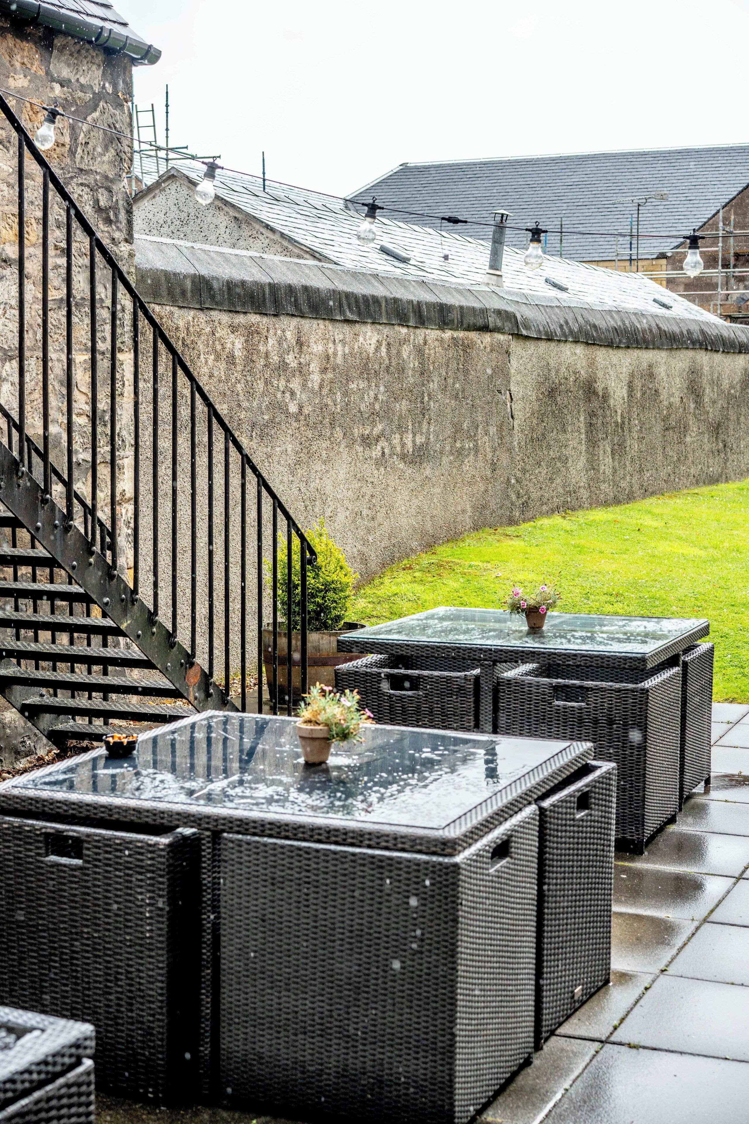 Mueble de mimbre exterior en un patio mojado con una pared de piedra y una escalera.
