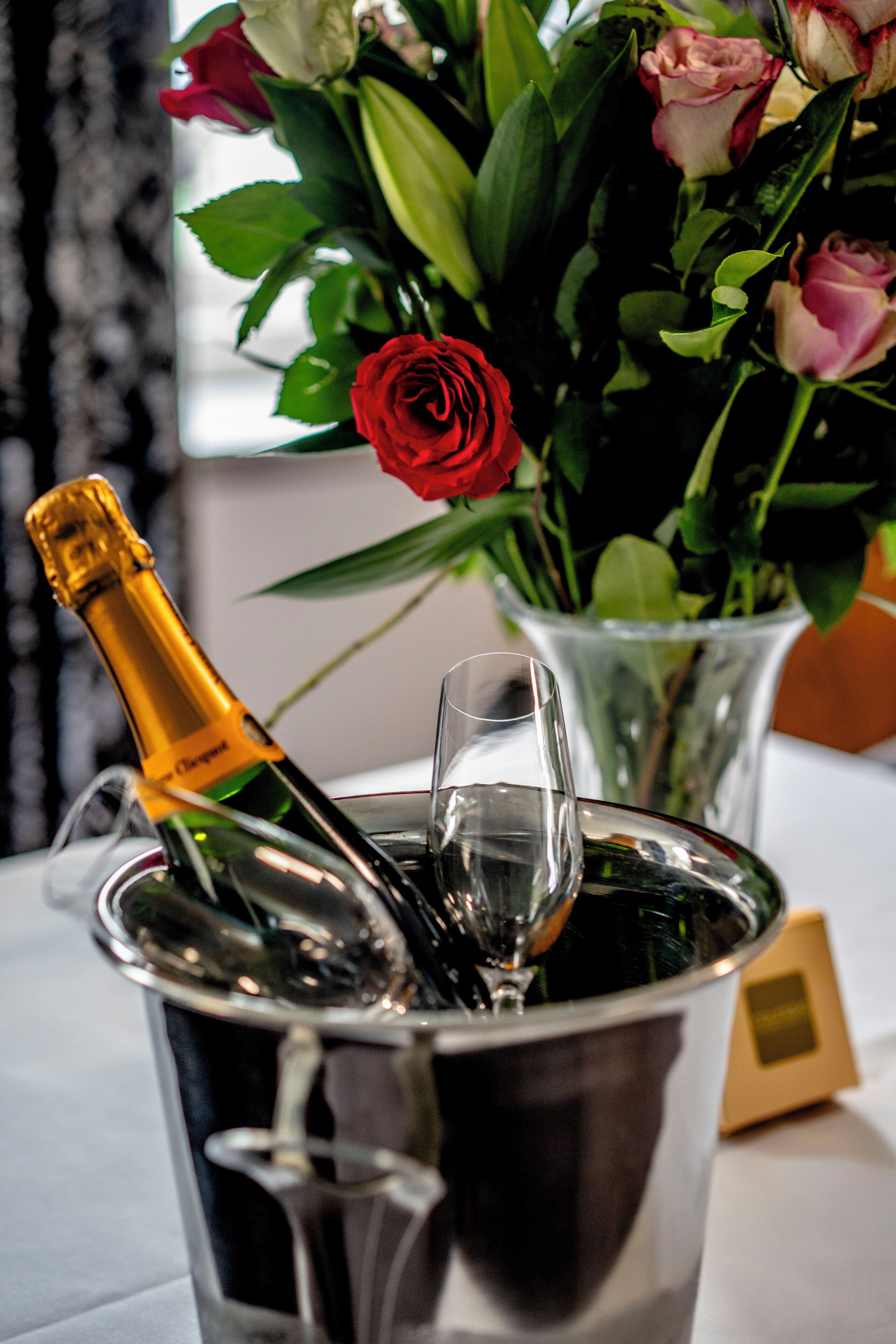 En flaske champagne i en metalspand sammen med et glas, en vase med blomster og et lille indrammet billede på et bord.