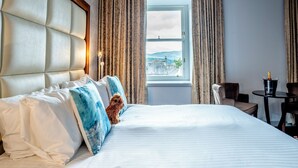 A hotel room with a large bed, a view of the outdoors, a small table with a bottle, and a chair. - Colessio (Stirling)