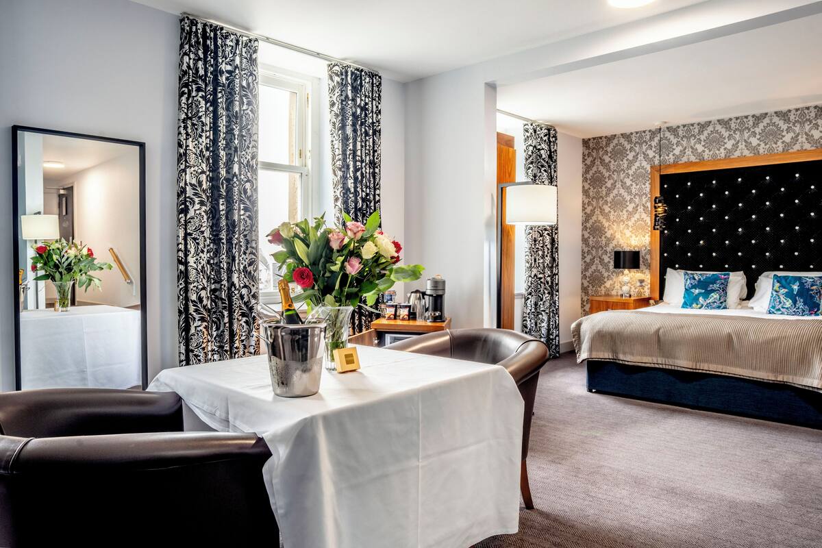 Une chambre d’hôtel avec un lit, une table à manger ornée d’un vase de fleurs, une chaise, un miroir et une lampe.