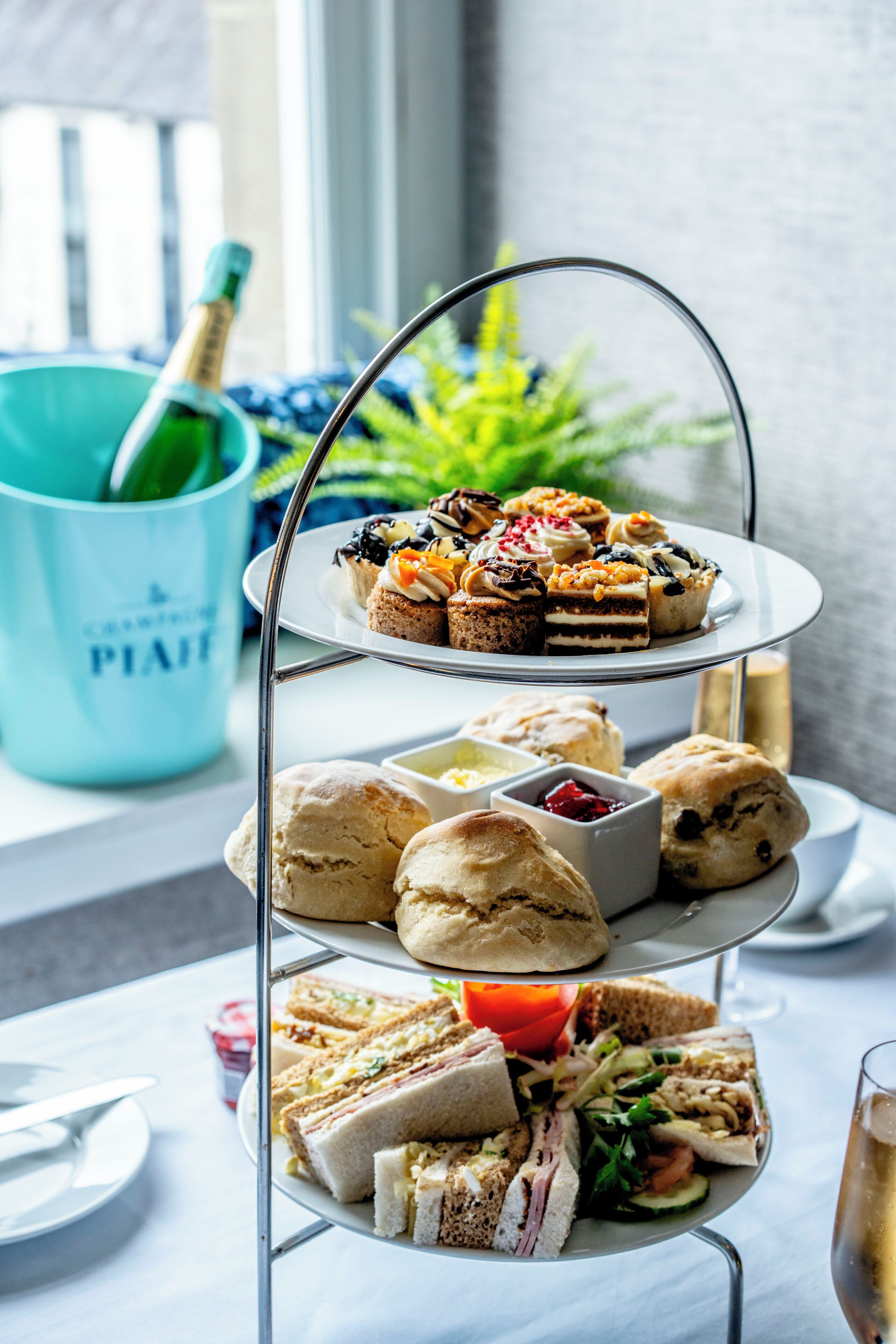 A tiered dessert and sandwich display with a champagne bottle and a blue cup in the background.