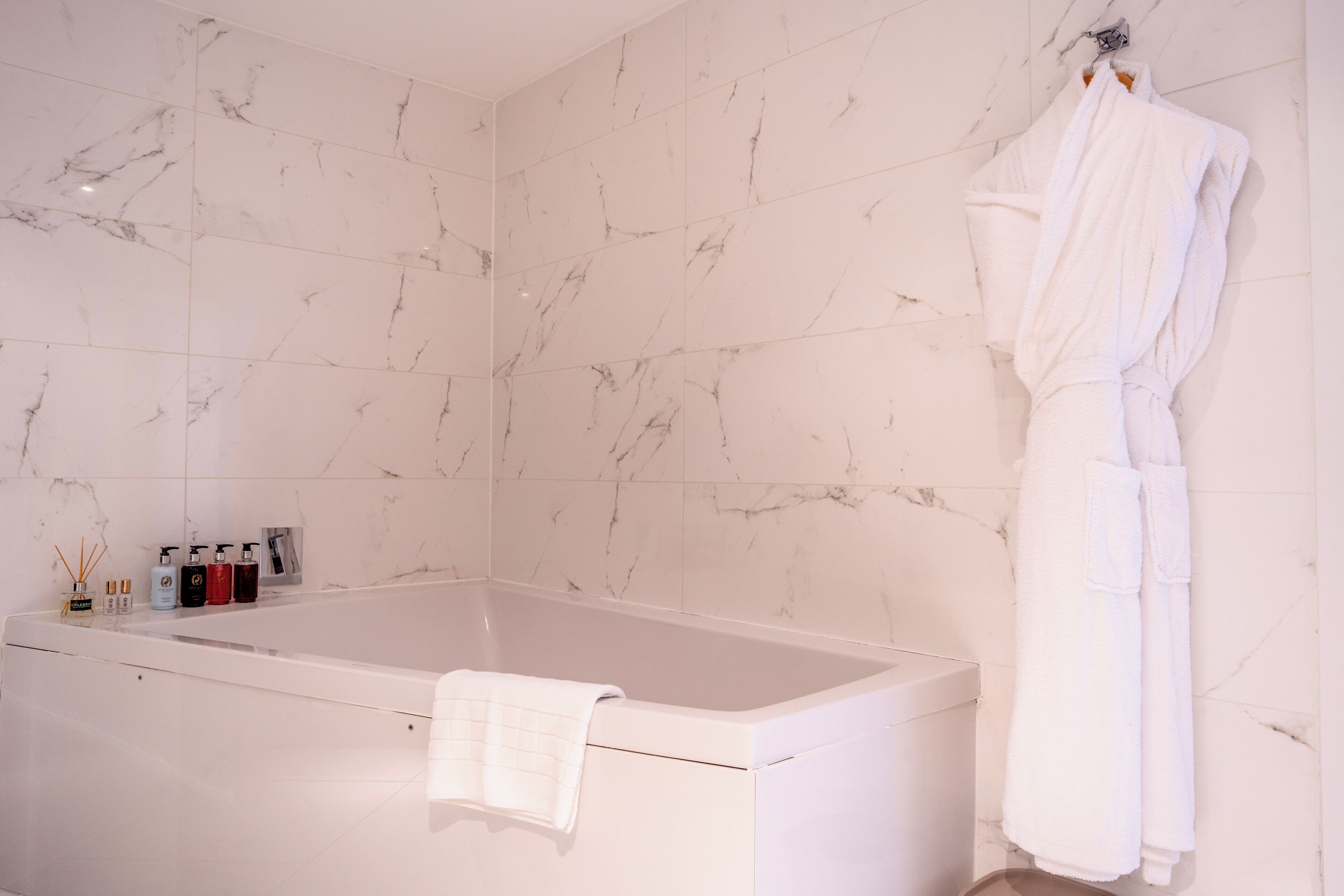 A bathroom with a marble-tiled wall, a bathtub, a white robe hanging on the wall, and a towel on the tub.