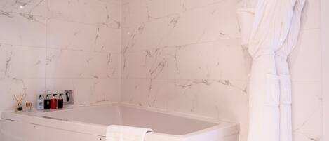 A bathroom with a marble-tiled wall, a bathtub, a white robe hanging on the wall, and a towel on the tub.