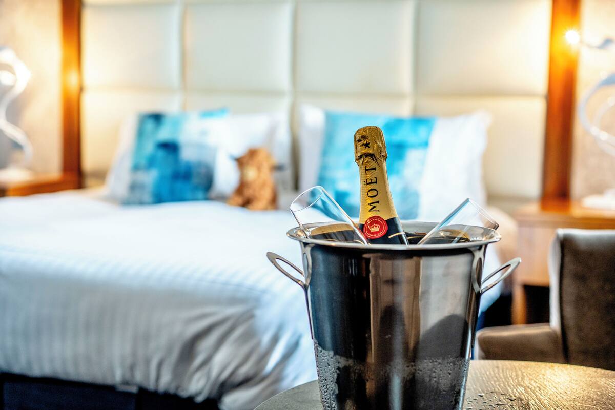 Un mini-bar contenant une bouteille de champagne Moët, un verre et une cuillère. Le mini-bar se trouve dans une chambre d’hôtel équipée d’un lit, d’oreillers et d’une table de chevet avec une lampe.