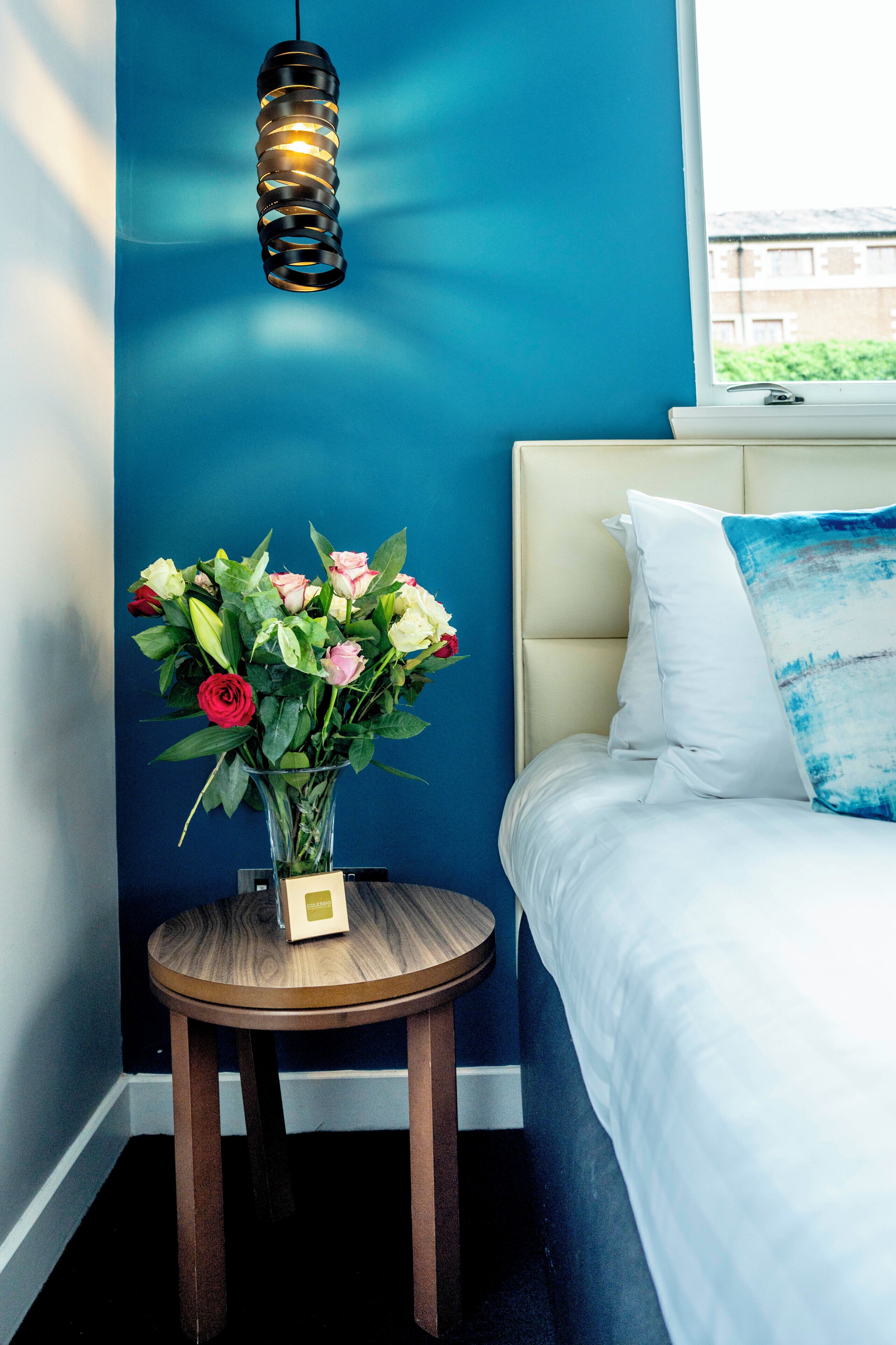 A bedroom with a bed, a bedside table with a vase of flowers, and a hanging light fixture.