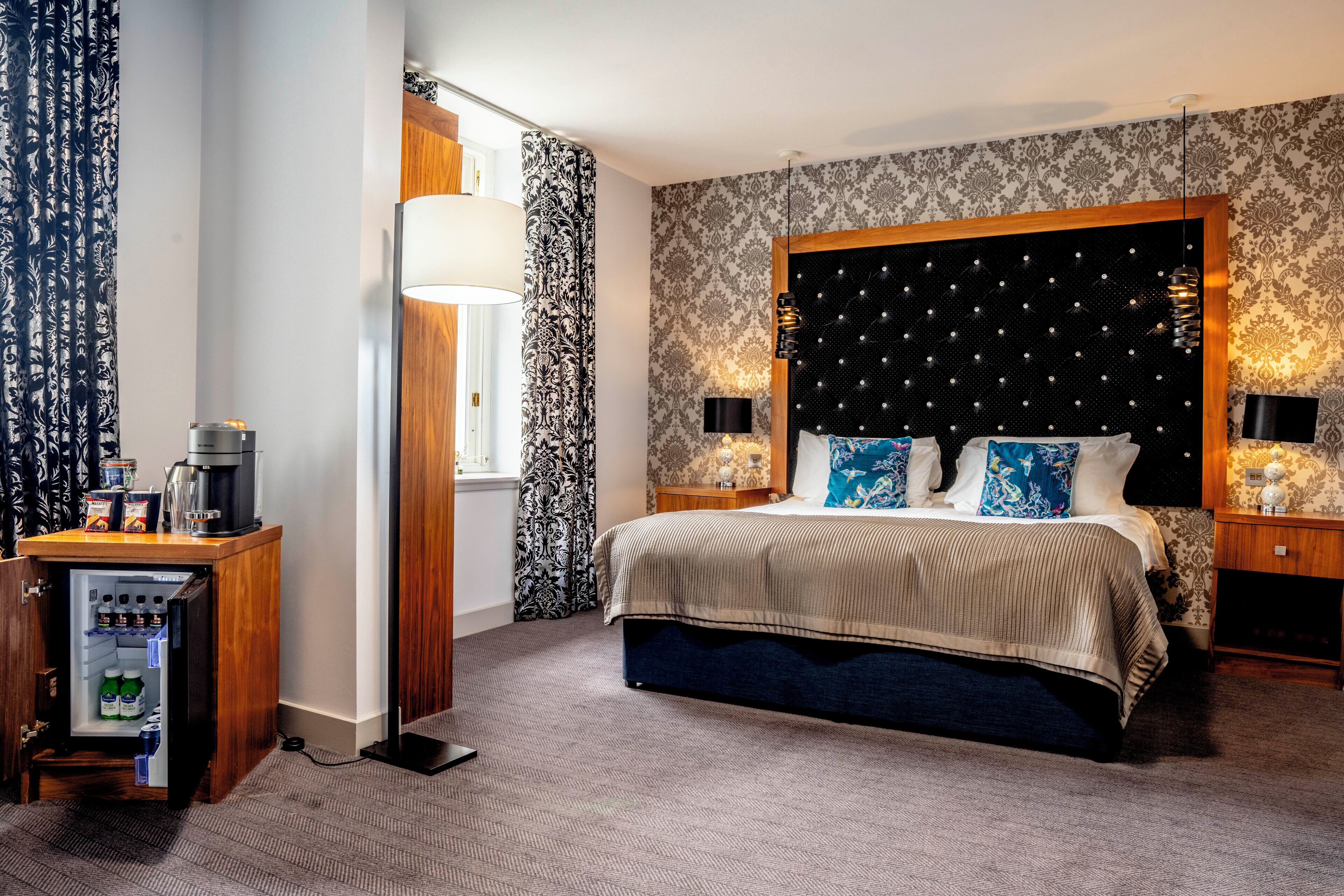 Una habitación de hotel con una cama grande, un minibar, una lámpara de pie y papel tapiz estampado.