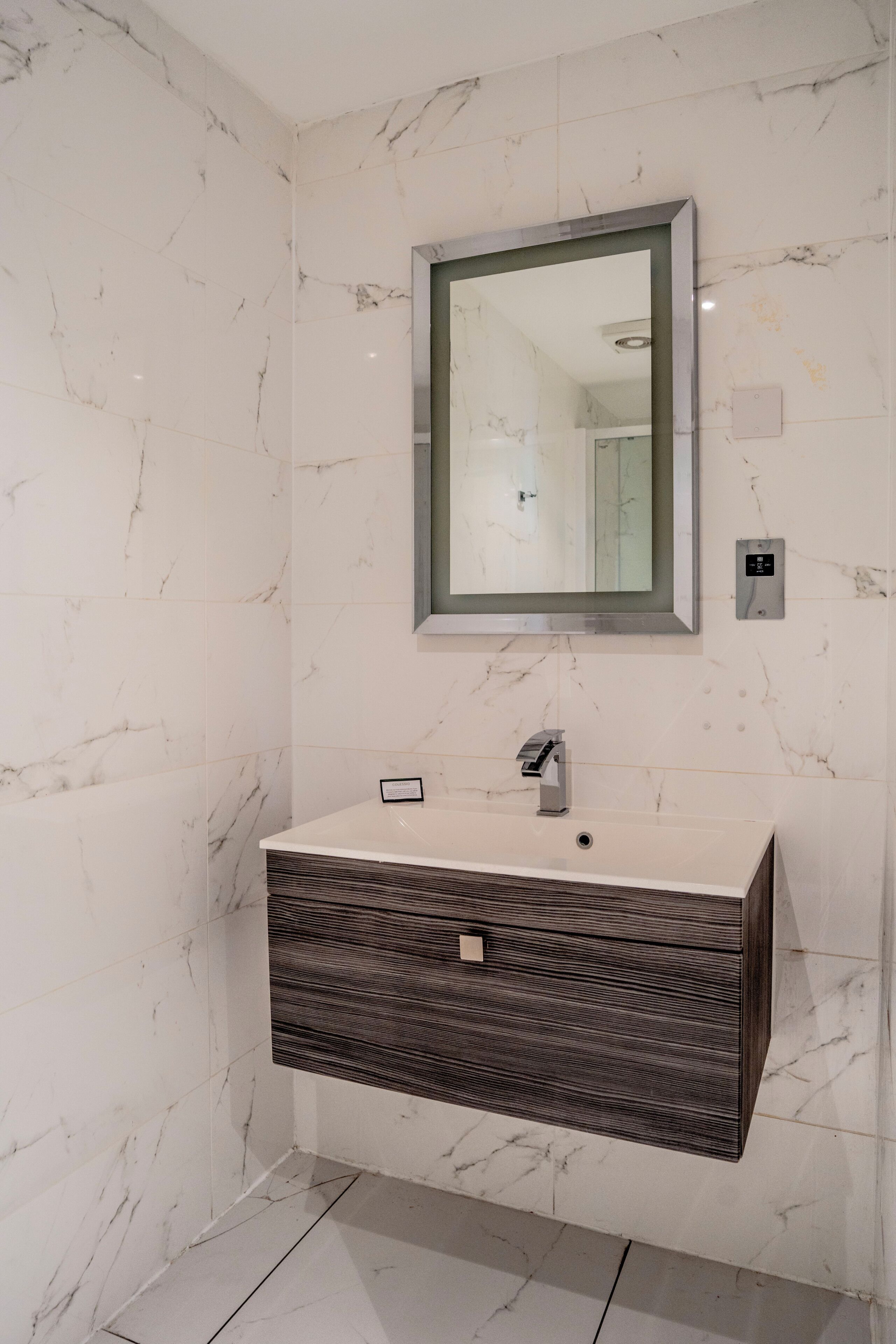 Een badkamer met een marmeren wand, een rechthoekige spiegel, een modern wastafelmeubel met een donker houten lade en een lichtschakelaar aan de muur.