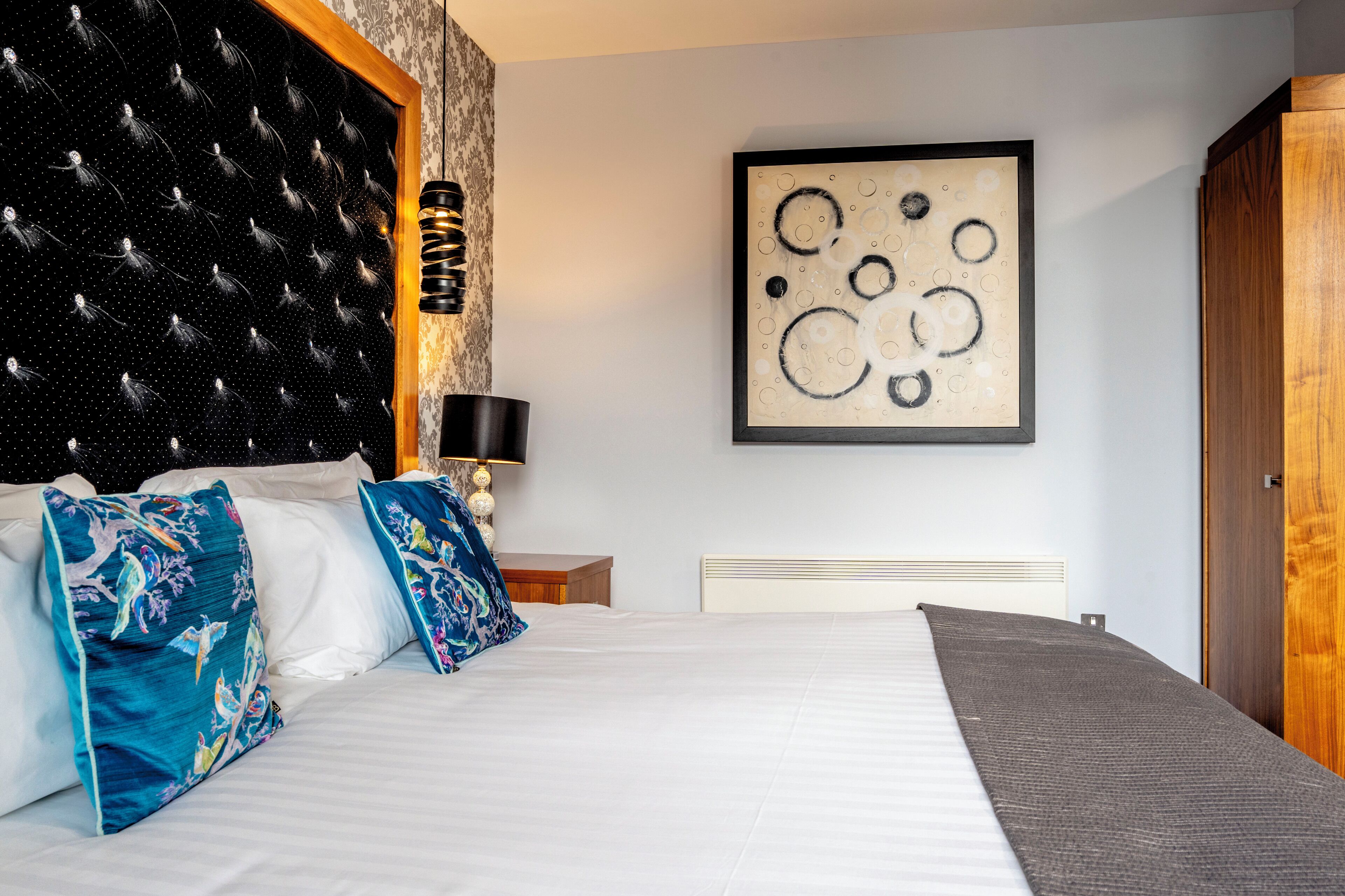 A hotel room with a large bed, a headboard with a tufted design, a framed abstract artwork on the wall, and a wooden door.