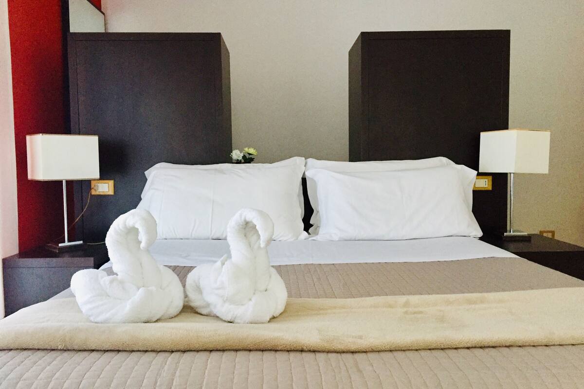 A hotel room with a bed, two bedside lamps, and two white swan-shaped pillows.