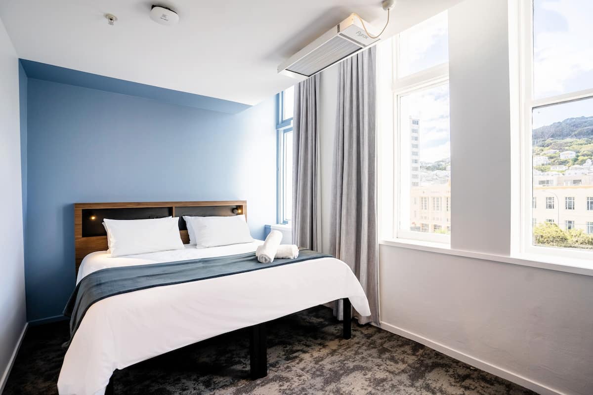 Een hotelkamer met een bed, een raam met gordijnen, een plafondlamp en uitzicht op gebouwen.