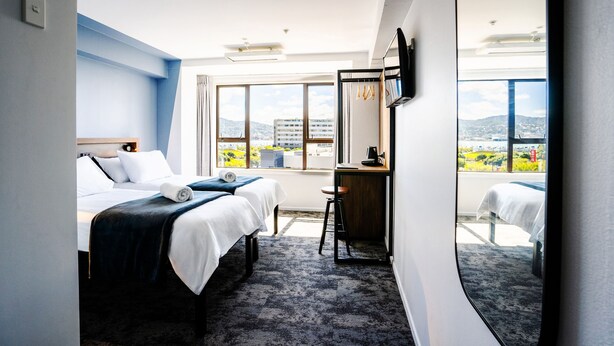 A hotel room with two beds, a desk, a chair, and a large window offering a city view. - Haka House Wellington (Wellington)