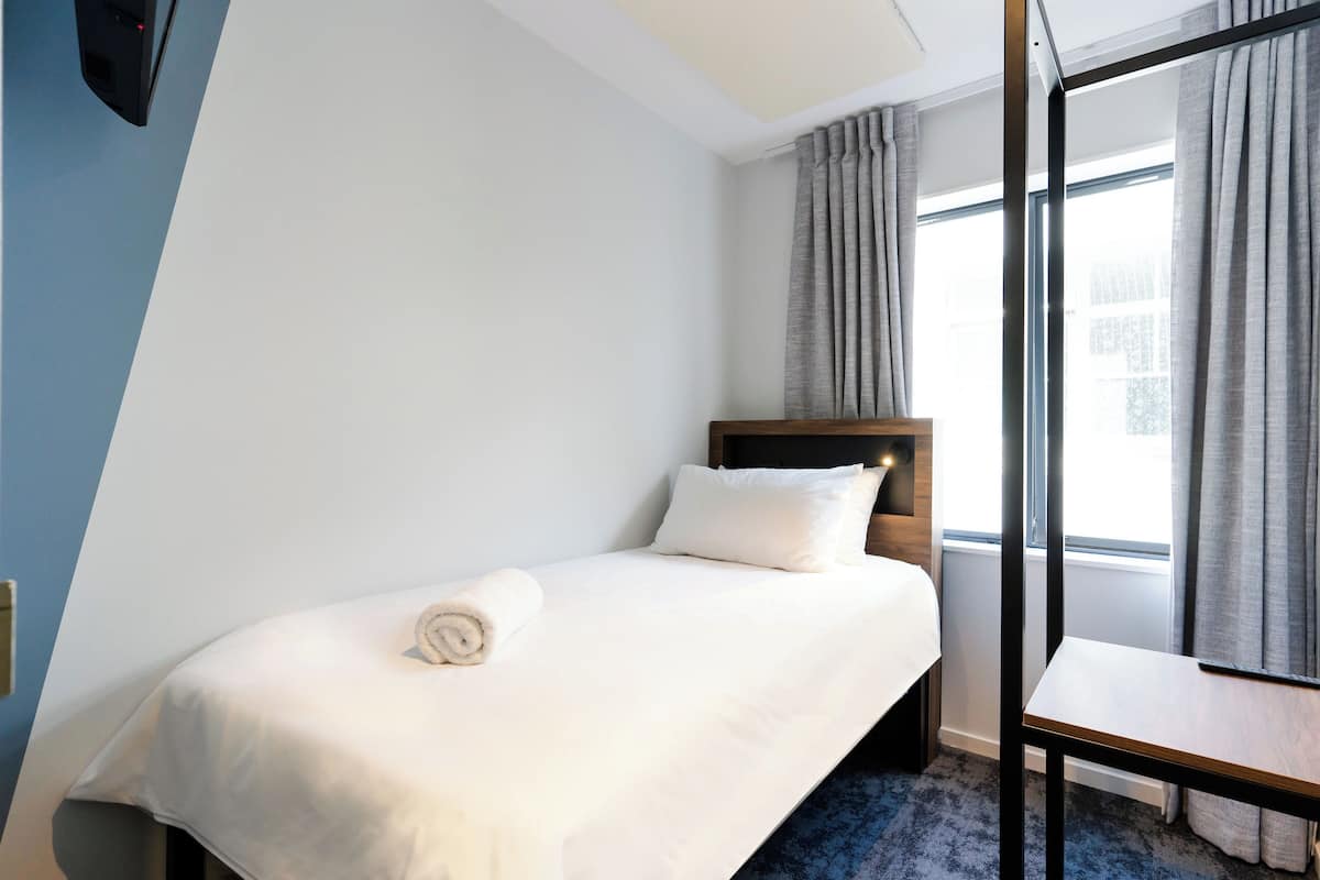Een hotelkamer met een net opgemaakt bed, een houten nachtkastje, een stoel en een raam met gordijnen.