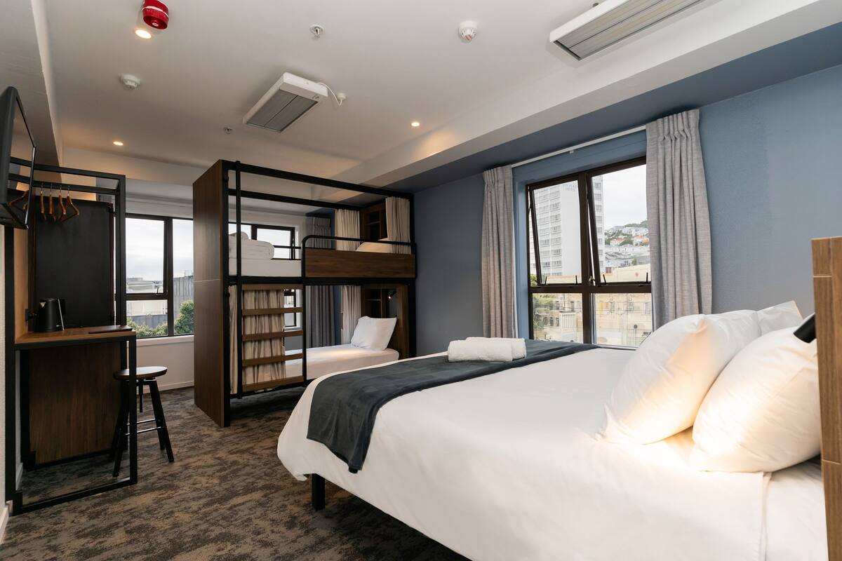 Een moderne hotelkamer met een groot bed, een bureau met stoel, een stapelbed en uitzicht op de stad door het raam.