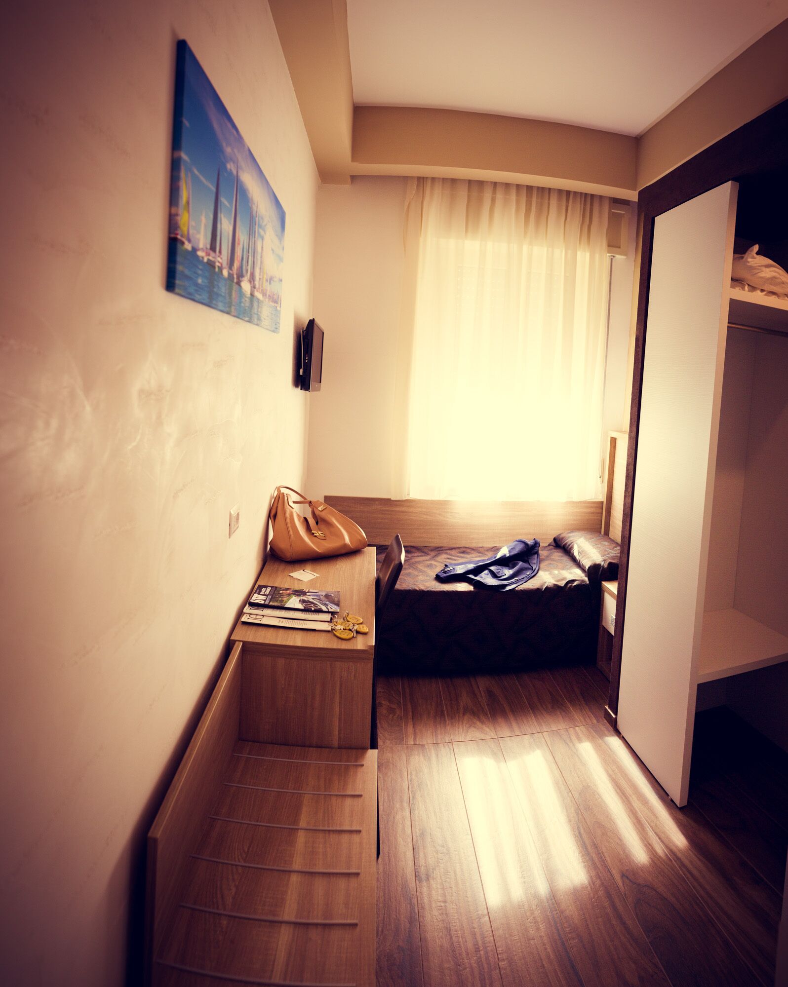 Una habitación de hotel pequeña con un banco de madera, una cama individual, un armario y un cuadro en la pared.