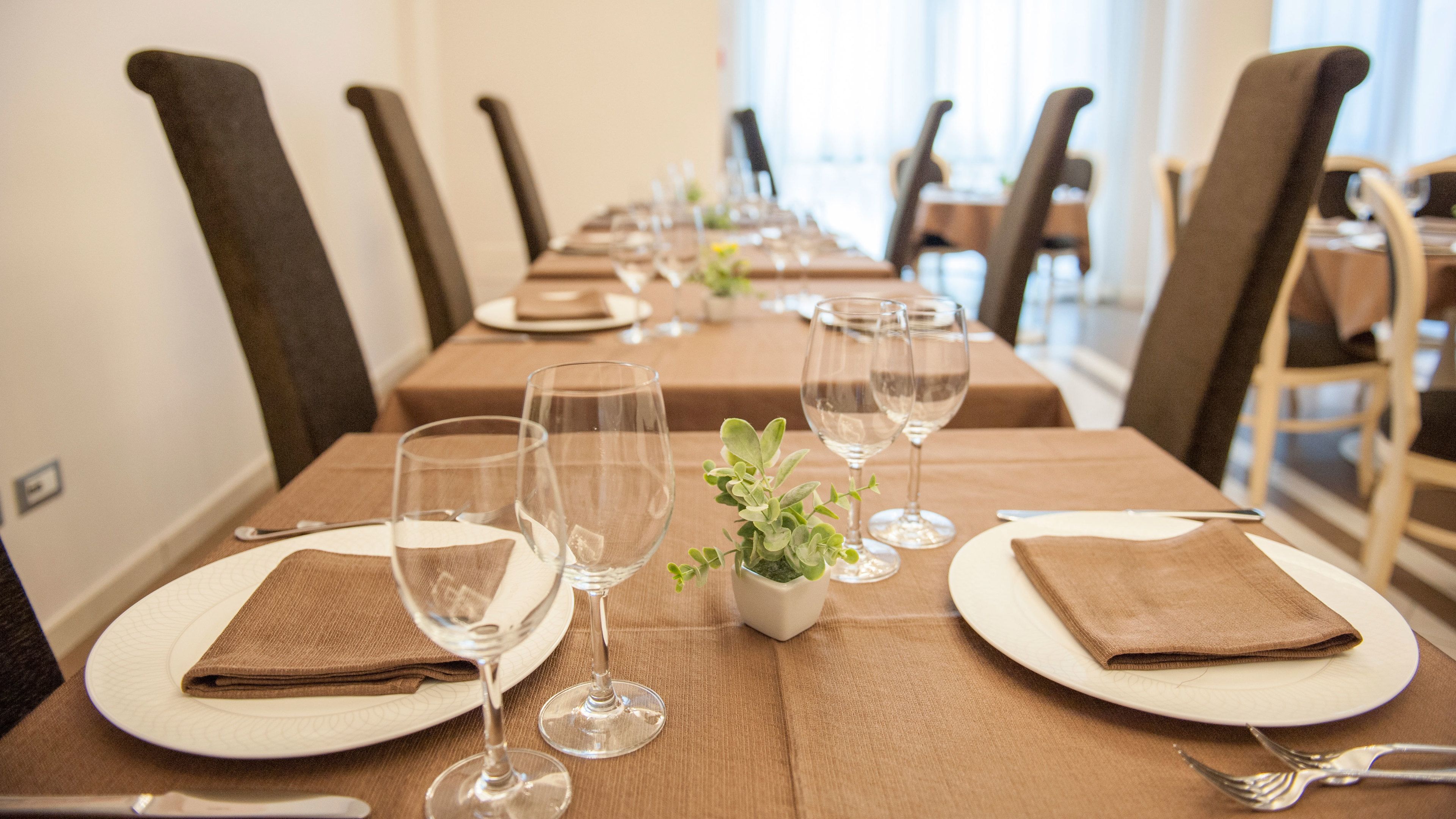 Una larga mesa de comedor con múltiples sillas, dispuesta con platos blancos, servilletas marrones dobladas y copas de vino transparentes.