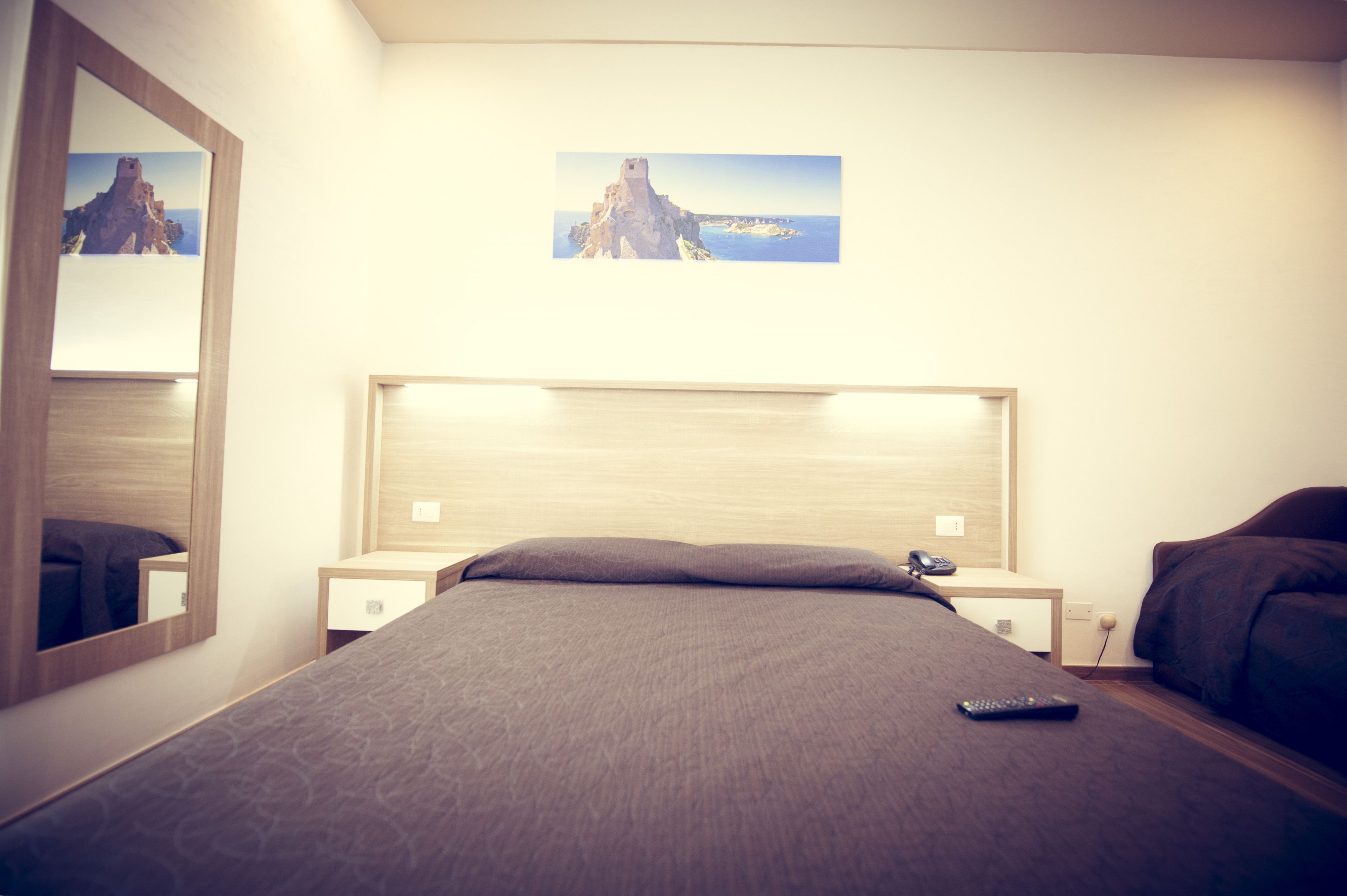 Ein Hotelzimmer mit Bett, Nachttisch, Spiegel und einem Wandbild mit einer Küstenlandschaft.