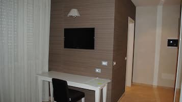 A hotel room with a white desk, a black chair, a flat-screen TV mounted on the wall, a lamp, and a door leading to another room.