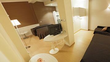 A modern hotel room with a bed, a desk with a computer, a small round table with a lamp, and a bathroom visible through an open door.