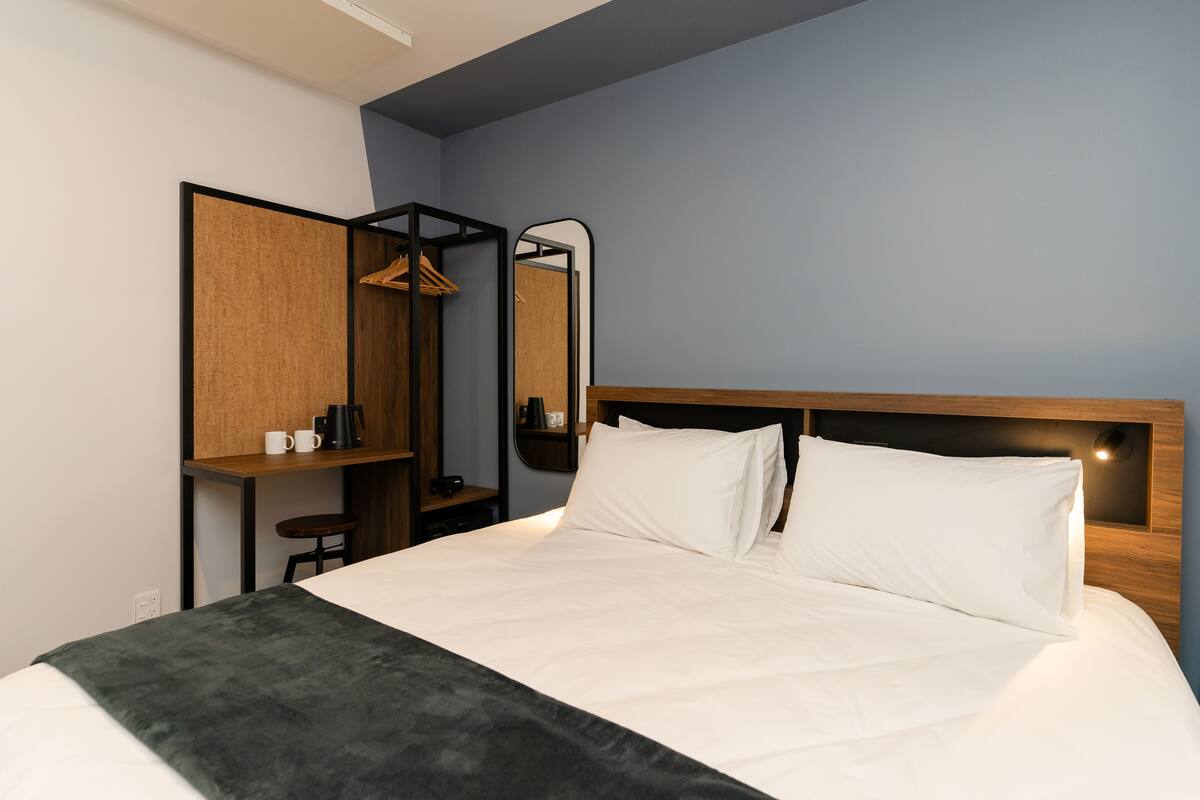 A modern hotel room with a neatly made bed, a wooden headboard, a small desk with a chair, a mirror, and a wardrobe.