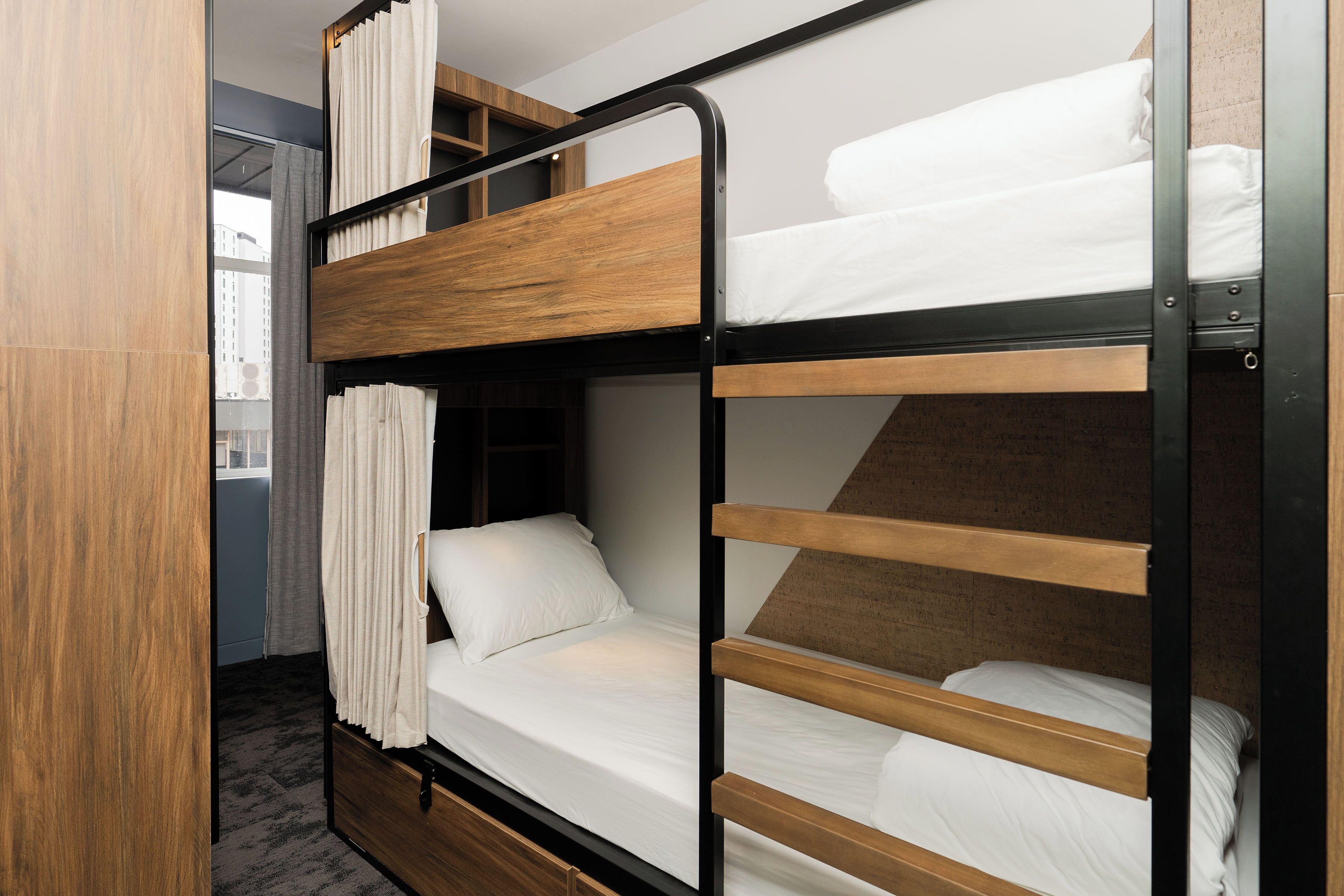 A bunk bed room with a wooden headboard, a window with curtains, and a view of the city.