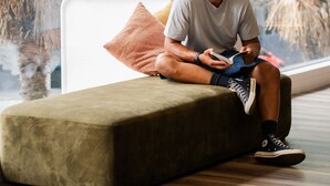 A person sitting on a green sofa in a room with large windows, a white wall, and a hanging light fixture. - Haka House Auckland City (Auckland)