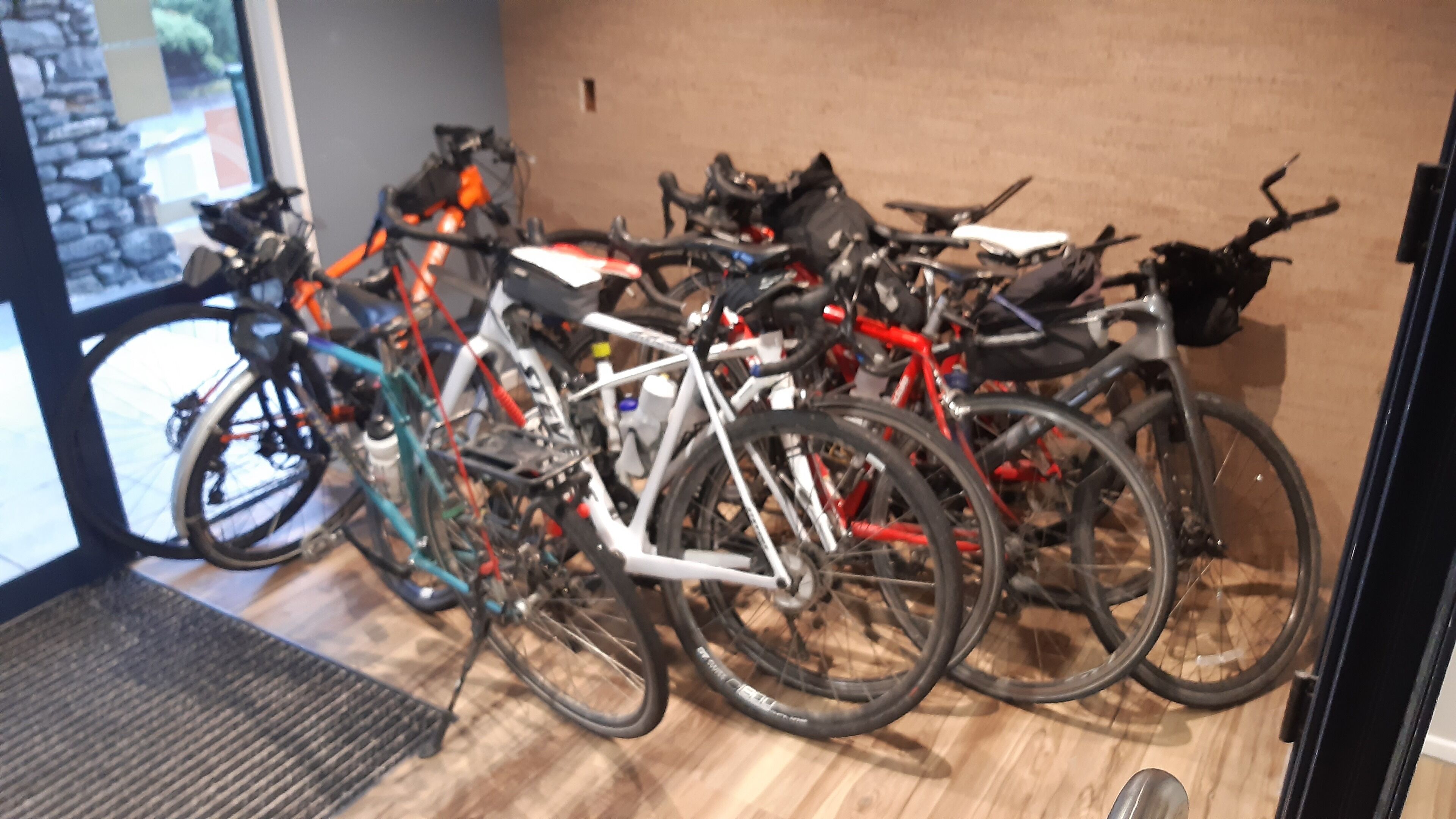 A row of bicycles parked indoors, with a wooden floor and a glass door leading outside.