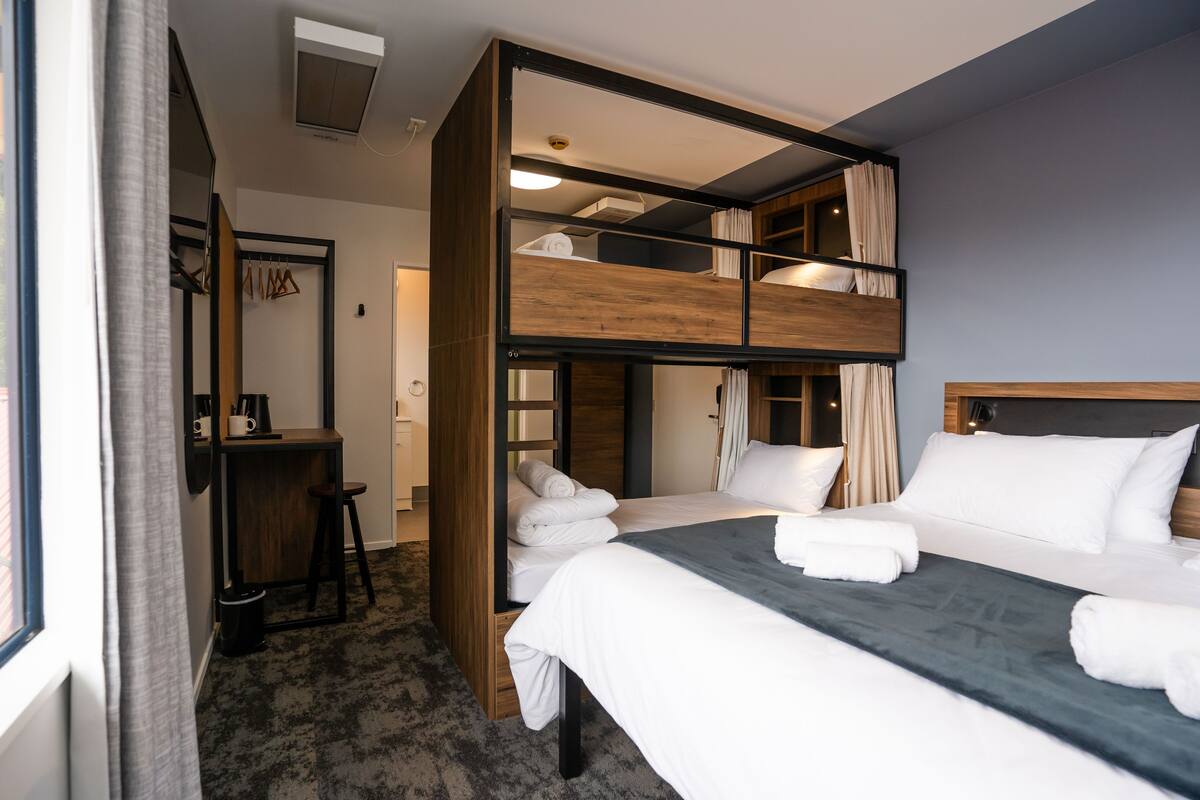 A hotel room with a bunk bed, a desk, and a large window.