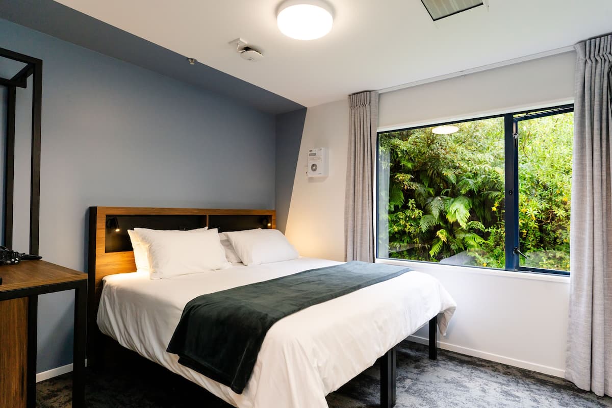 A hotel room with a bed, a desk, a window with curtains, and a view of greenery outside.
