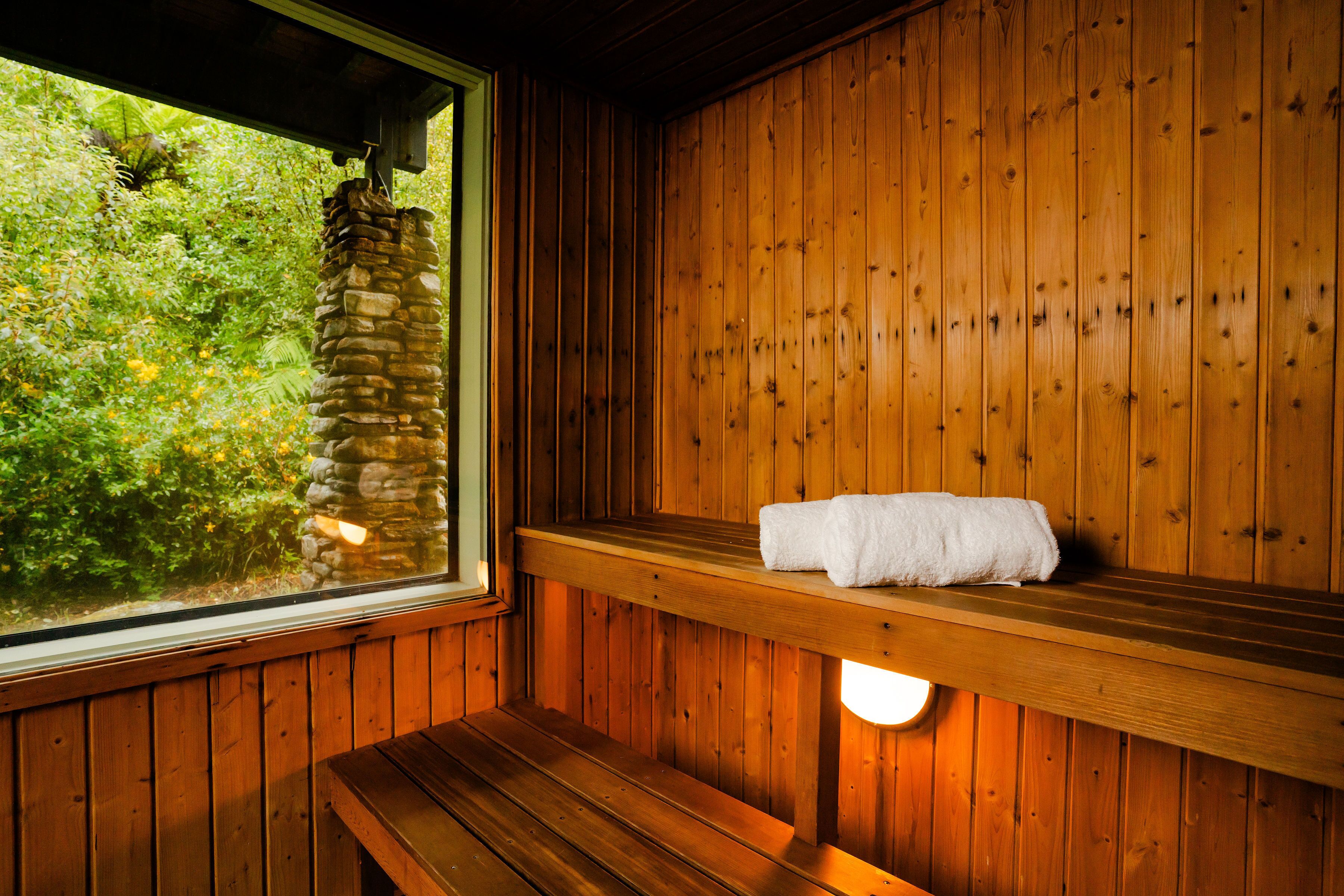 A wooden sauna with a bench and a towel on a shelf, a stone pillar, and a window with a view of greenery.