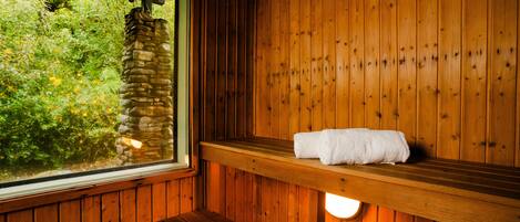 A wooden sauna with a bench and a towel on a shelf, a stone pillar, and a window with a view of greenery.