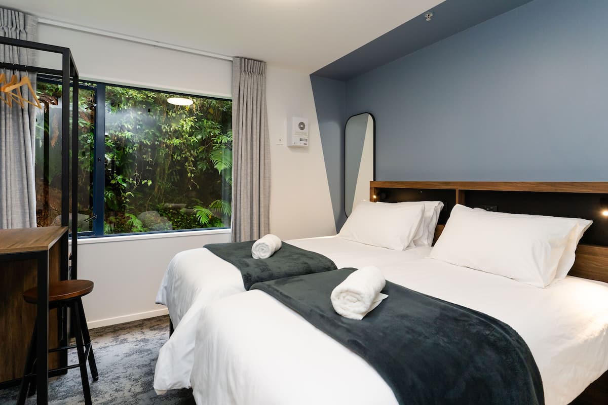 A hotel room with two beds, a desk, and a large window with a view of greenery.