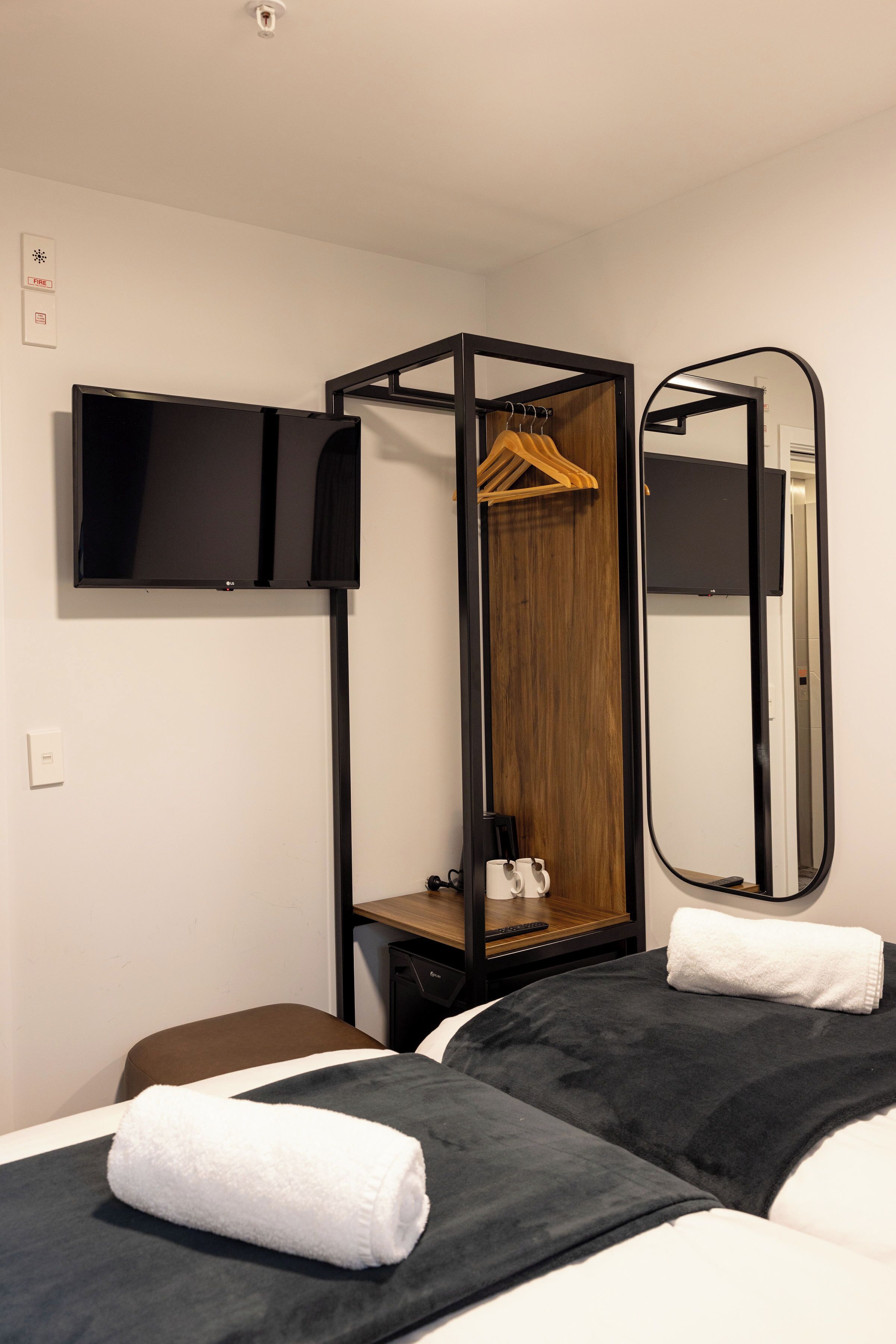 A hotel room with a bed, a TV mounted on the wall, a wooden wardrobe, a mirror, and a towel on the bed.