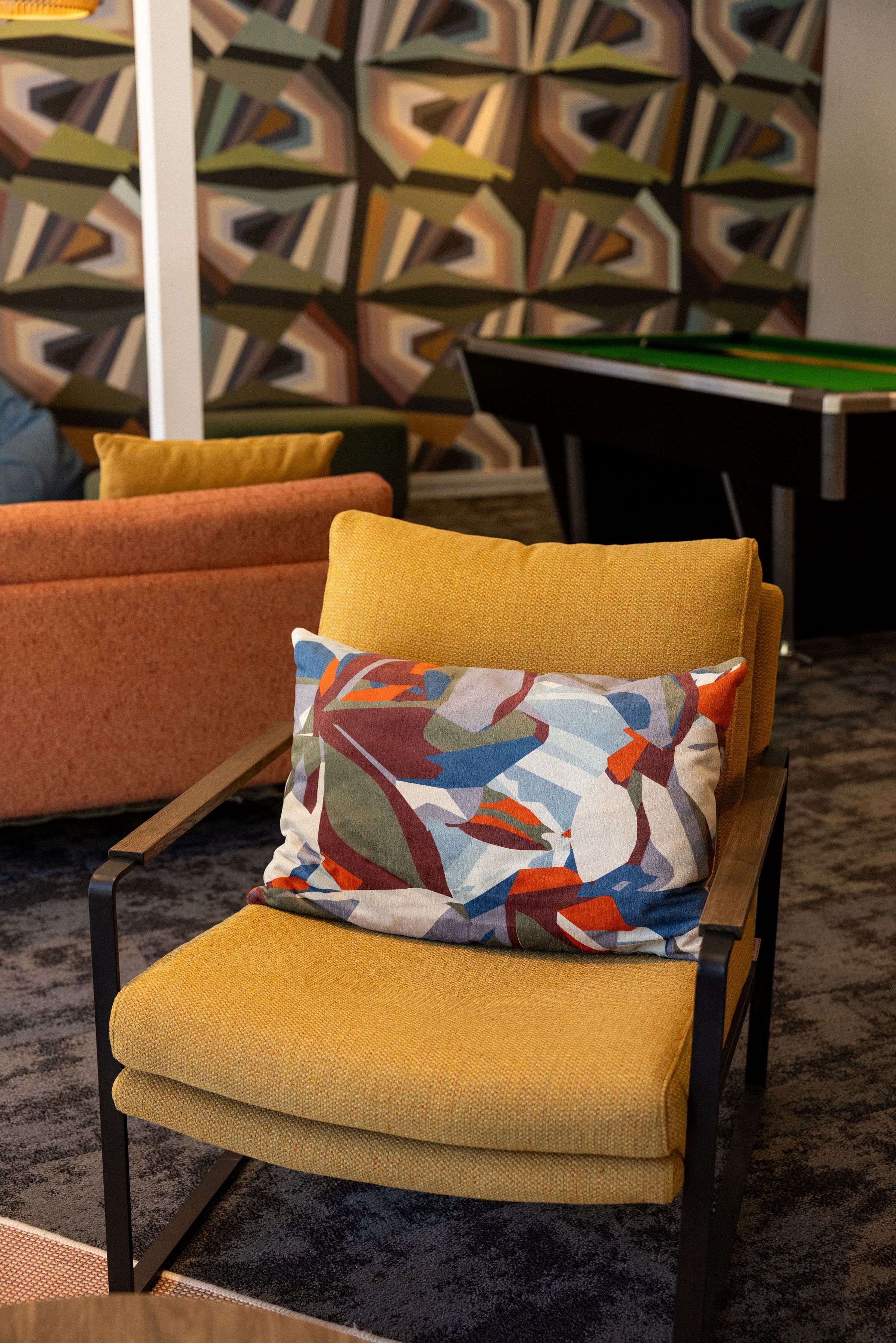 Un área de descanso moderna con un sillón amarillo y un cojín colorido, una mesa de billar y un papel tapiz estampado al fondo.