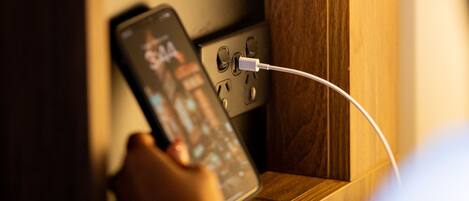 A person holding a smartphone with a charging cable plugged in, near a wooden cabinet with a black object on top.