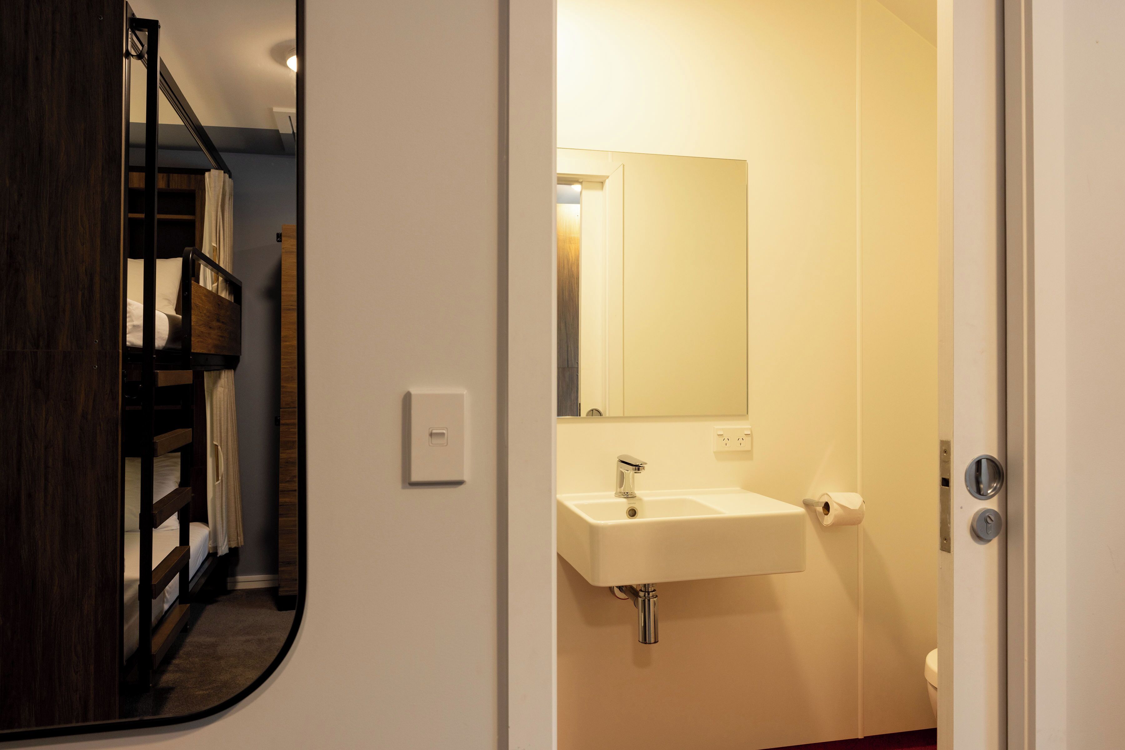 Une salle de bain avec un lavabo blanc, un miroir et des toilettes. On peut apercevoir un lit superposé en bois à travers une porte ouverte.