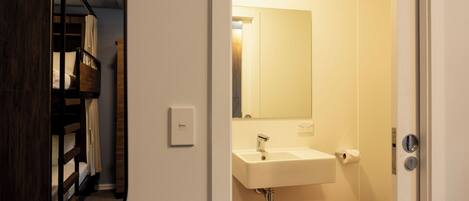 Une salle de bain avec un lavabo blanc, un miroir et des toilettes. On peut apercevoir un lit superposé en bois à travers une porte ouverte.