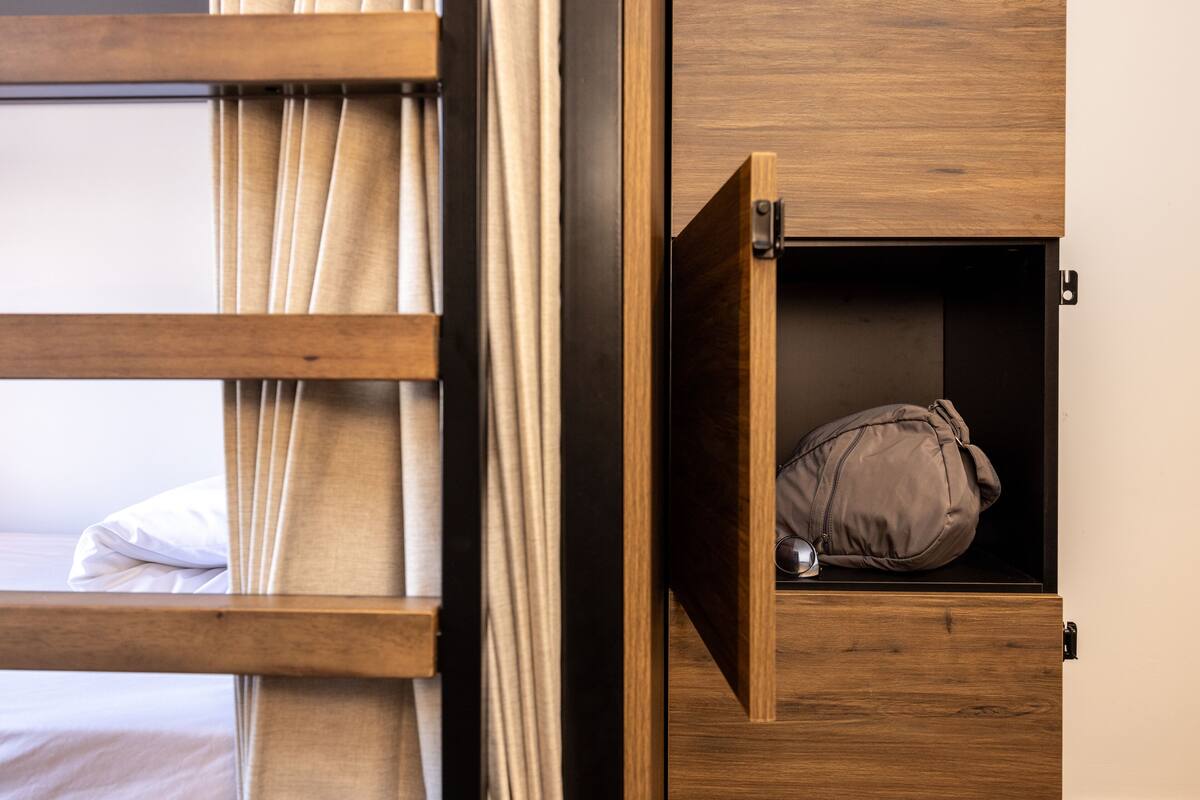 Una cama litera con estructura de madera, un mueble cajón extraíble con un almohadón dentro, y una cortina que cubre parcialmente la ventana.
