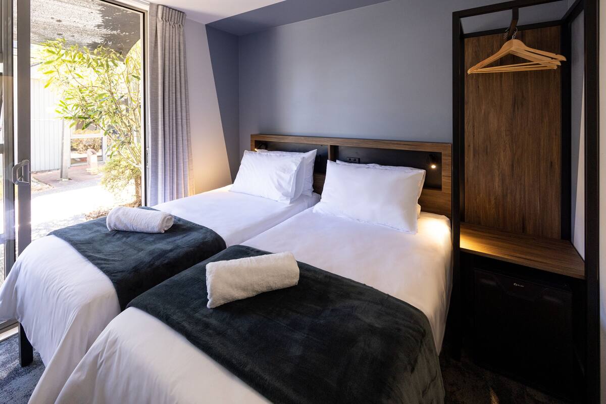 Habitación de hotel con dos camas, cabecero de madera, perchero y vista al exterior.