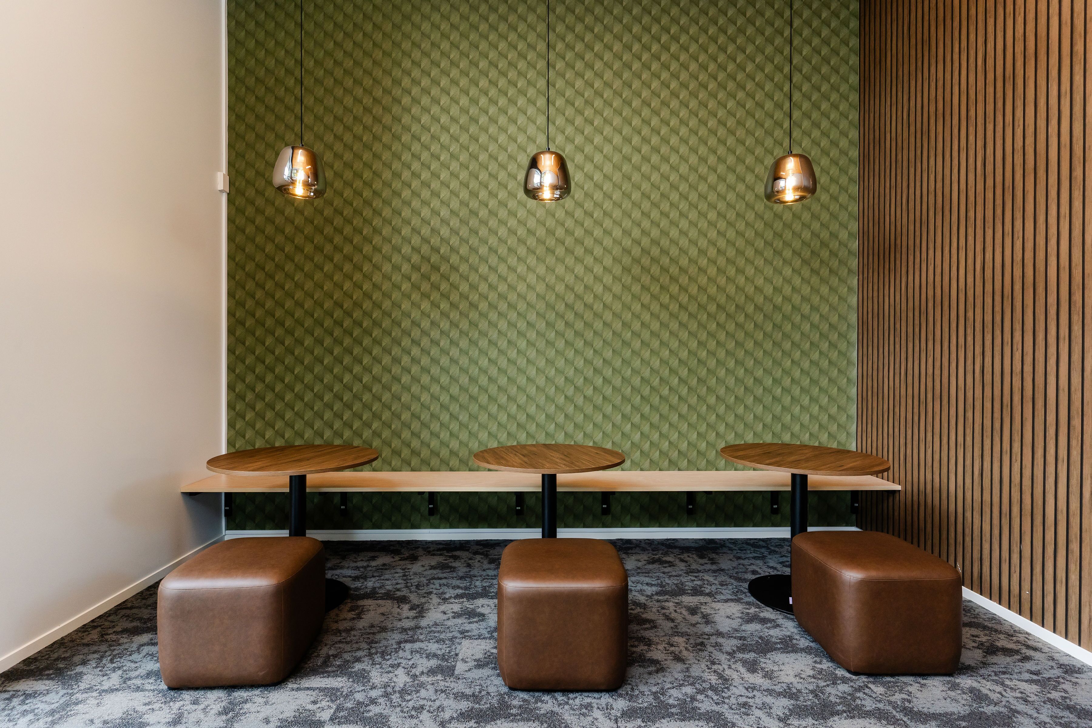 A seating area with three wooden tables and three brown ottomans, a green patterned wall, and hanging pendant lights.