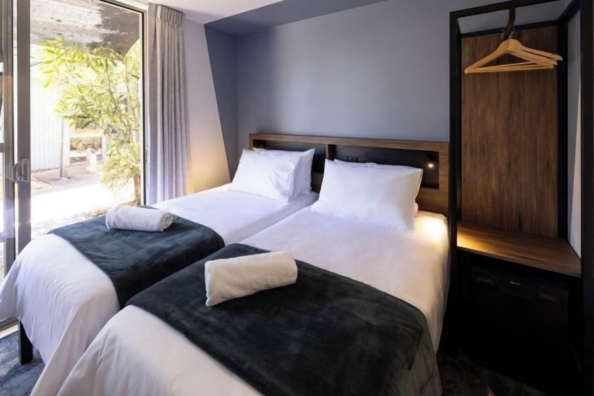 A hotel room with two beds, a wooden headboard, a hanging coat rack, and a view of greenery outside.