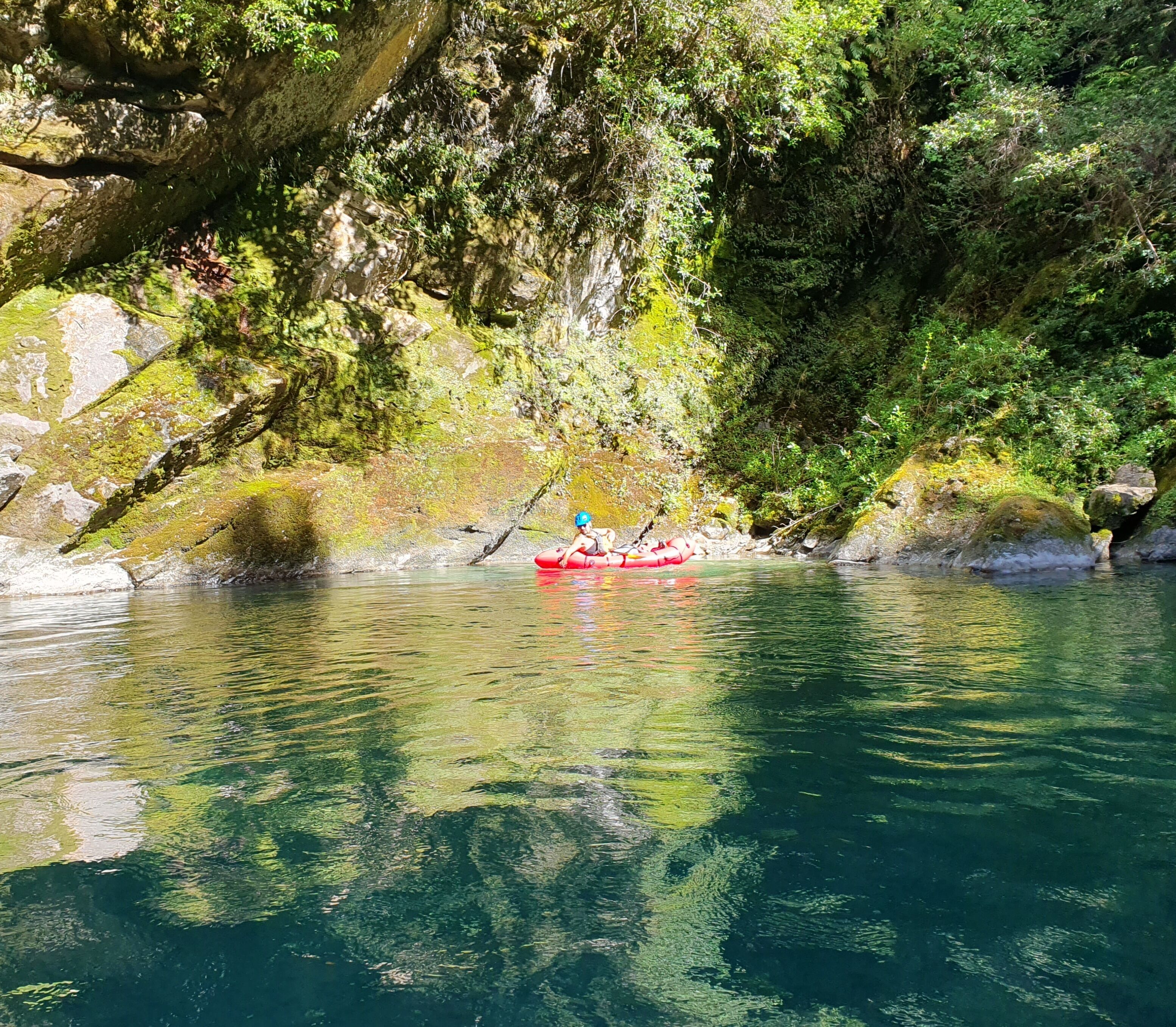 Un río tranquilo con un kayak rojo y una persona remando, rodeado de exuberante vegetación y acantilados rocosos.
