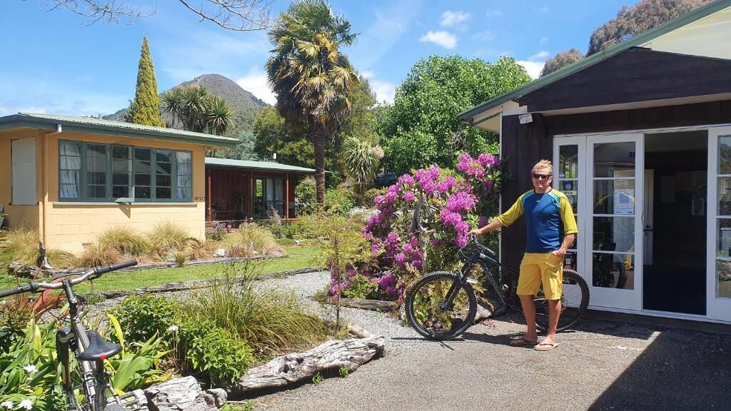 A building with a door and windows, surrounded by a garden with purple flowers and a bicycle.