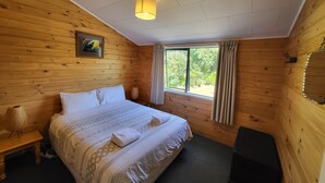 A wooden cabin room with a bed, a window with curtains, a ceiling light, and a painting on the wall. - Off The Beaten Trail Accommodation & Bike Tours (Murchison)