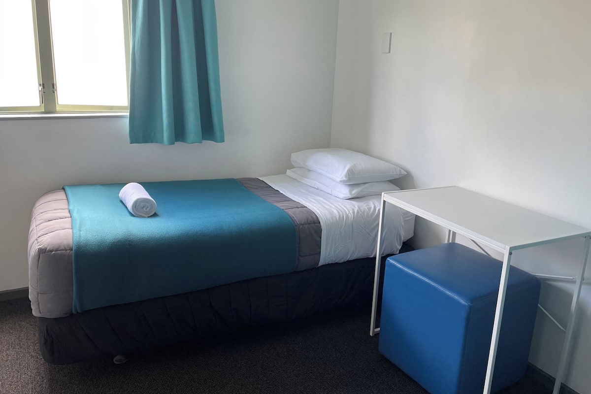 A single bed with a teal blanket and a rolled towel, a white pillow, a small white table, and a blue bench.