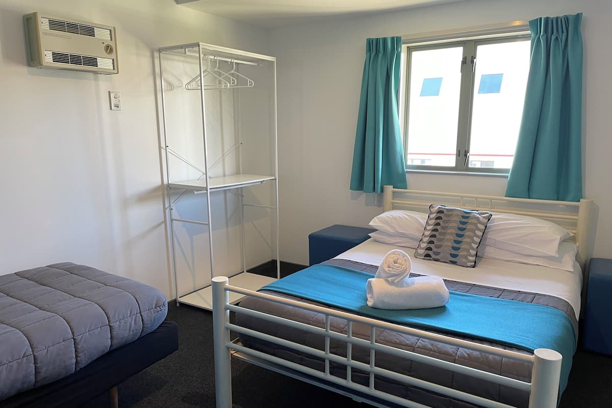 A single bed with a blue blanket, a white pillow, and a rolled towel. There is a white air conditioning unit on the wall, a metal drying rack, and a window with teal curtains.