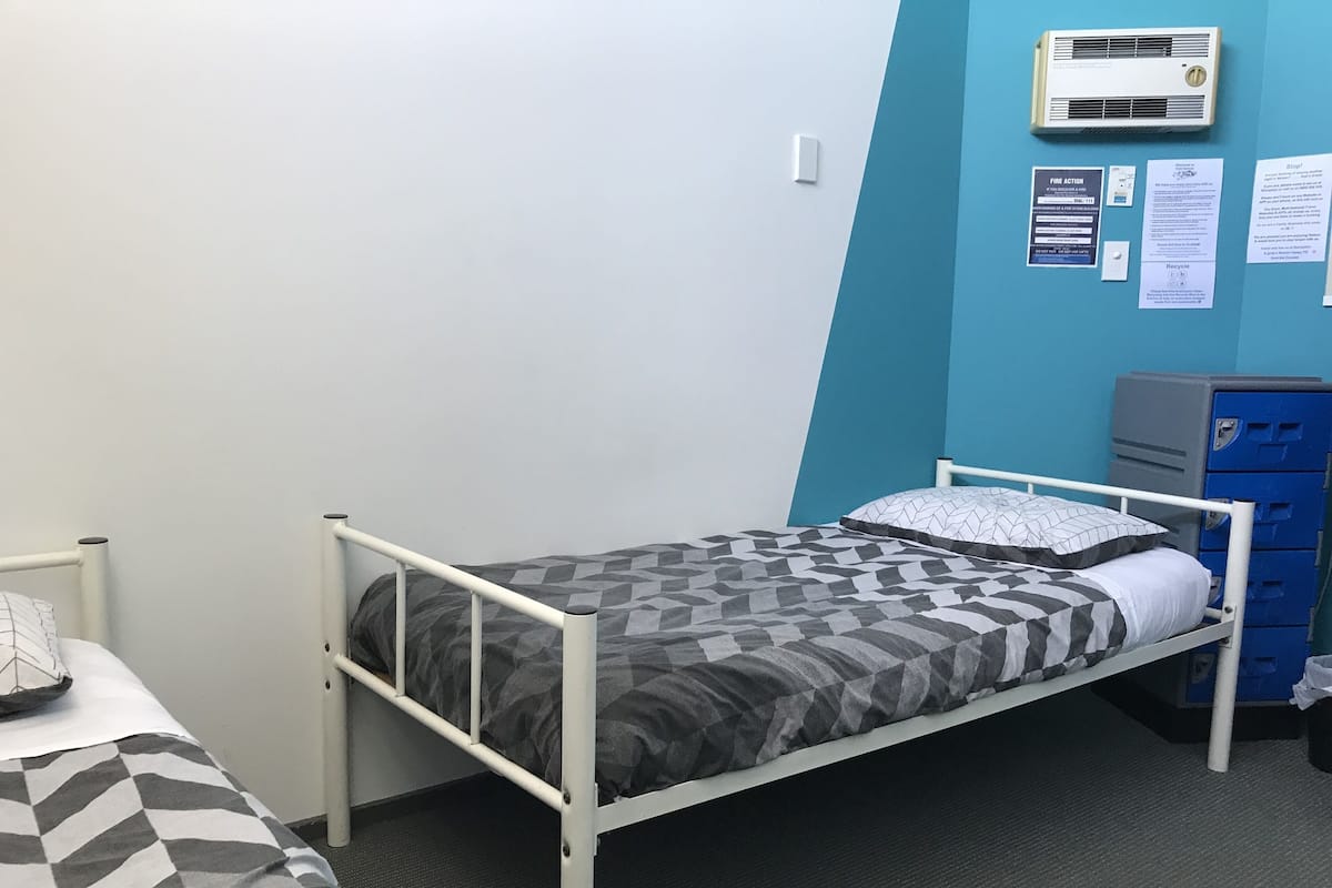 A single-bed room with a metal frame, grey patterned bedding, a white pillow, and a blue wall with informational posters.
