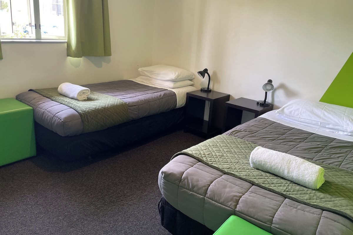 A hotel room with two beds, a green chair, a nightstand with a lamp, and a window with curtains.