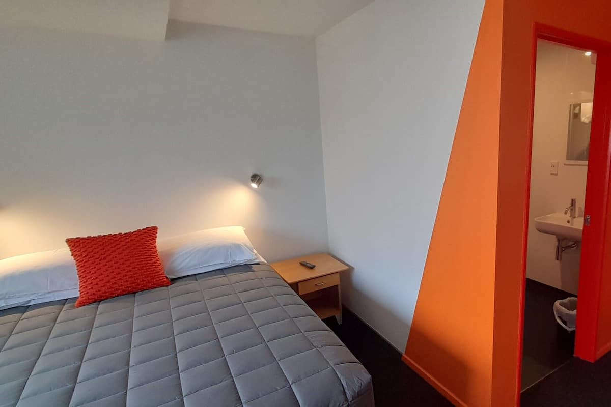 A hotel room with a bed, a nightstand, a red door, and a bathroom visible through an open door.