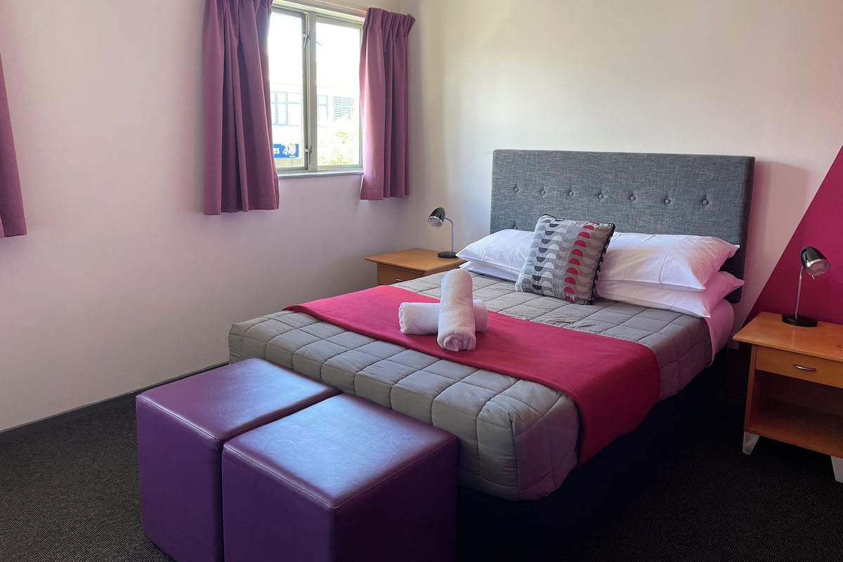 A neatly arranged hotel room with a bed, a bedside table, a nightstand, a window with curtains, and a small purple ottoman.